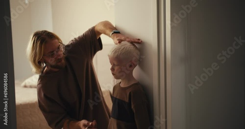 A little albino boy with white hair stands near the wall while his blond dad with a beard and glasses measures his height and then shows the boys height to him. Measure your childs growth photo