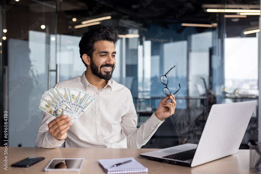 Happy young Muslim man sitting in office in front of laptop, talking on video call, showing and holding fan of money and glasses, bragging about achievement and success