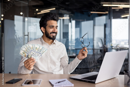 Happy young Muslim man sitting in office in front of laptop, talking on video call, showing and holding fan of money and glasses, bragging about achievement and success photo