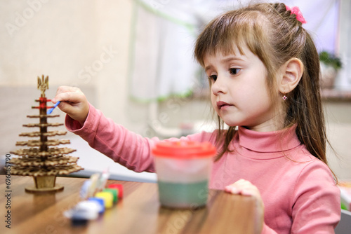 A little girl paints a wooden toy in the shape of a Christmas tree. She works on her masterpiece with concentration, carefully applying paint to each