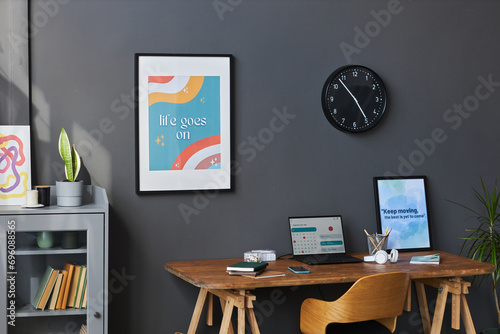 Part of students home office interior with wooden table and laptop, bookcase and motivational posters at dark-gray wall photo