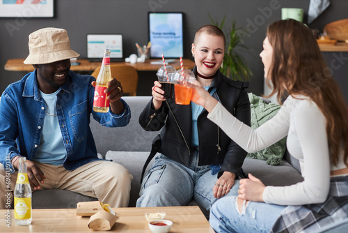 Medium full shot of three cheerful diverse young people toasting with soda while sitting on couch at home photo