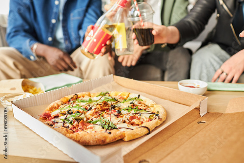 Close up shot of sauced pizza with greens on top in cardboard box on table, defocused peoples hands holding beverages in blurred background