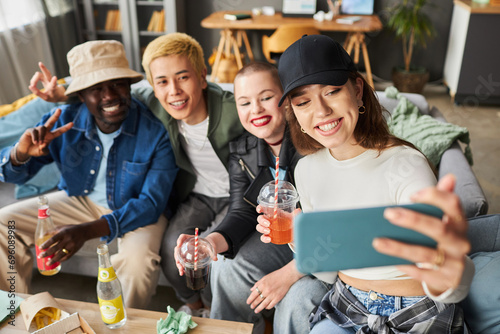 High angle shot of beaming multi-ethnic young people taking selfie during party in living room, focus on brunette girl in cap holding smartphone photo