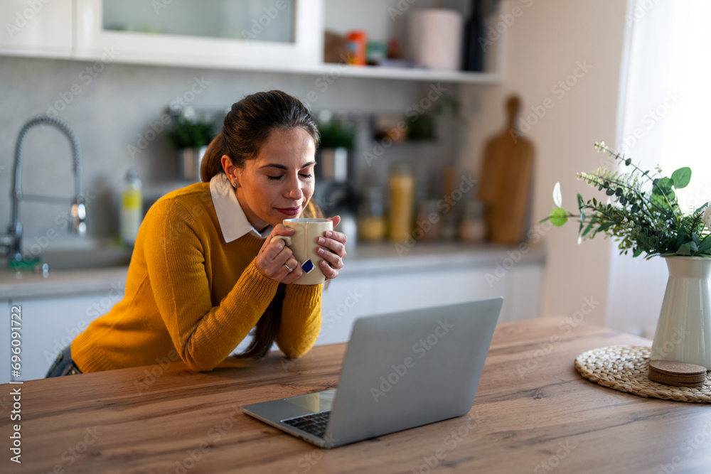 Smiling businesswoman having a break and drinking tea in front of laptop at desk. Young woman enjoying the smell of hot tea. - obrazy, fototapety, plakaty 
