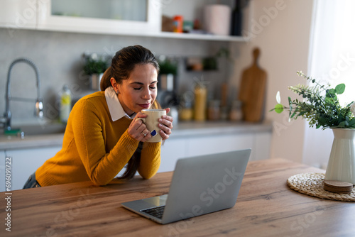 Smiling businesswoman having a break and drinking tea in front of laptop at desk. Young woman enjoying the smell of hot tea.