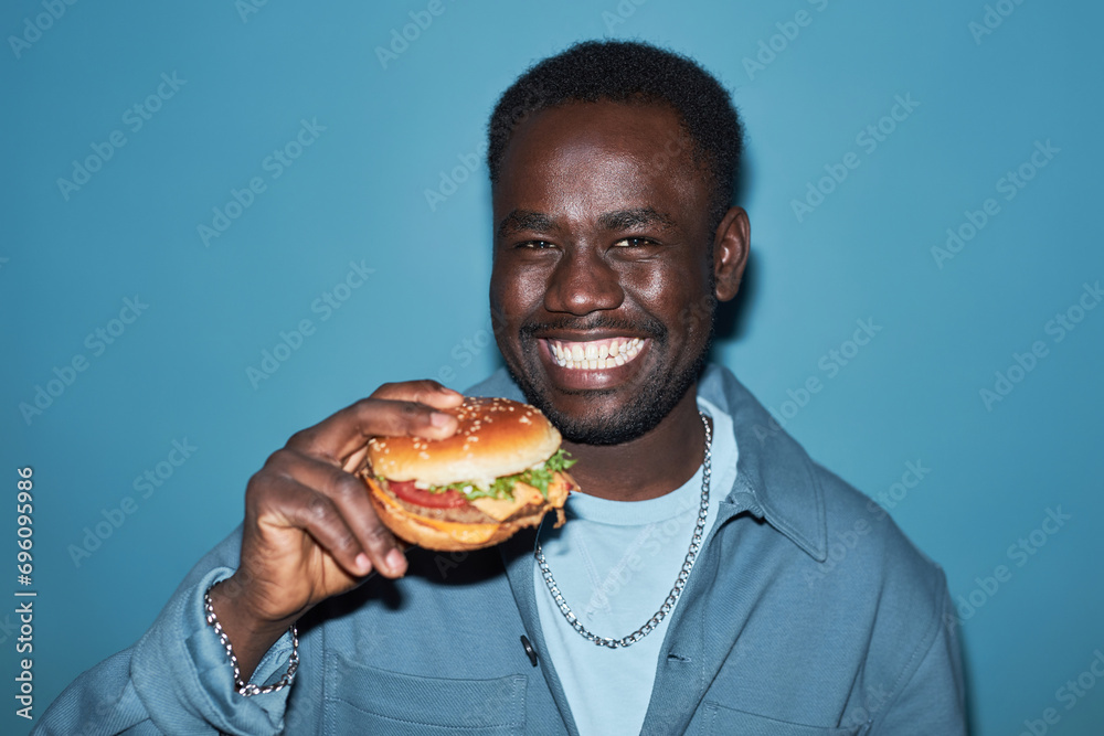 Medium close up shot of young African American man with toothy smile looking at camera and holding tasty burger while standing at light blue wall Medium close up shot of young African American man with toothy smile looking at camera and holding tasty burger while standing at light blue wall
