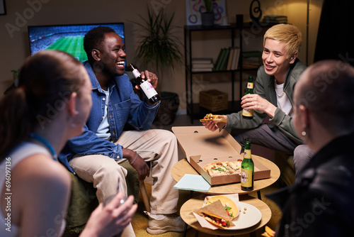 Medium full shot of diverse young adults sitting in living room laughing and talking while drinking and eating fast food