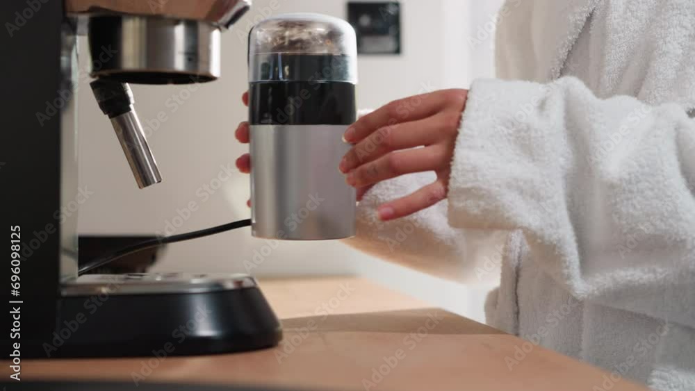 Woman grinds coffee beans in device at table closeup. Lady in terry robe mills grains in grinder at home. Processing of caffeine product to make drink