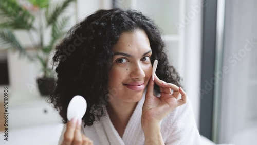 Close-up portrait of positive curly haired multinational woman in bathrobe with clean smooth flawless skin, removing make-up with cotton pads, spa, therapy, treatment, on background of bright bedroom. photo
