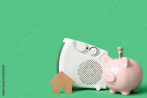 Electric heater with piggy bank, money and wooden house on green background. Concept of heating season