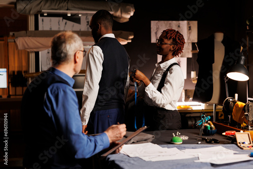 African american gentleman getting measurements taken by professional suitmakers in dark studio workspace. Masterful tailors looking for ideal client proportions for upcoming custom made suit photo