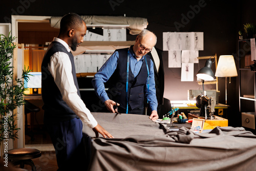 Meticulous elderly master tailor cutting fabric sheet for upcoming bespoke luxury fashion collection. Senior suitmaker showing african american apprentice expert craftmanship photo