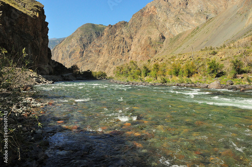 A shallow turbulent river with clear water flows through a deep canyon in the shadow of the setting sun from the mountains.