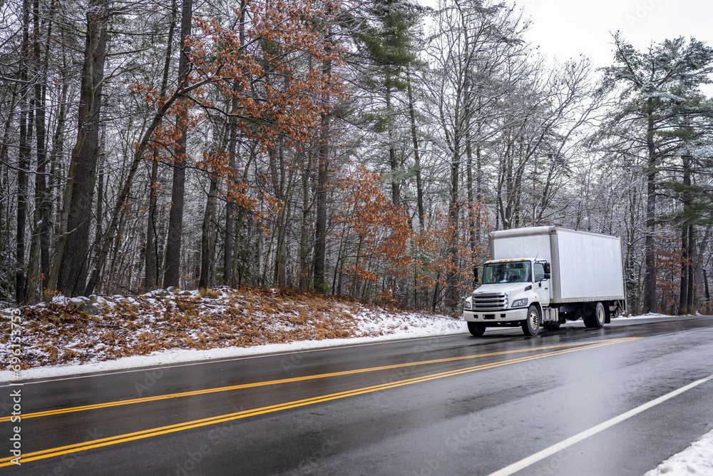 Middle duty white day cab rig semi truck with box trailer for local freight driving on the winter slippery road with snow and ice and snowy forest on the side