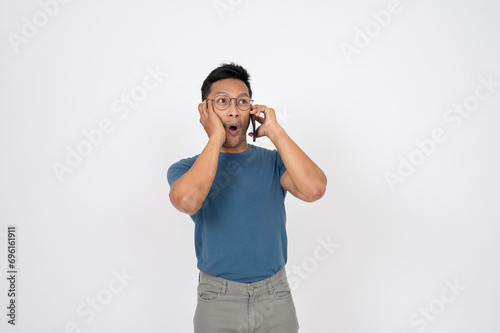 A surprised, shocked Asian man is talking on the phone with someone, isolated background. © bongkarn