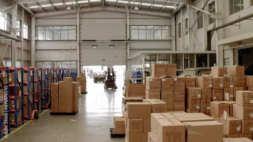 A forklift is parked between boxes at a warehouse of a Chinese factory. Hundreds of packaged products are organised on shelves at the storage facility Lots of sunlight is coming in through the windows photo