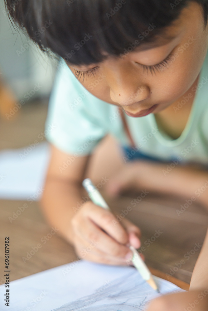 Close up portrait of Asian boy with black bangs, black eyes with a smiling face wearing a light green and lying on the floor of his house drawing a picture of a girl with braids. Education concept. Close up portrait of Asian boy with black bangs, black eyes with a smiling face wearing a light green and lying on the floor of his house drawing a picture of a girl with braids. Education concept.