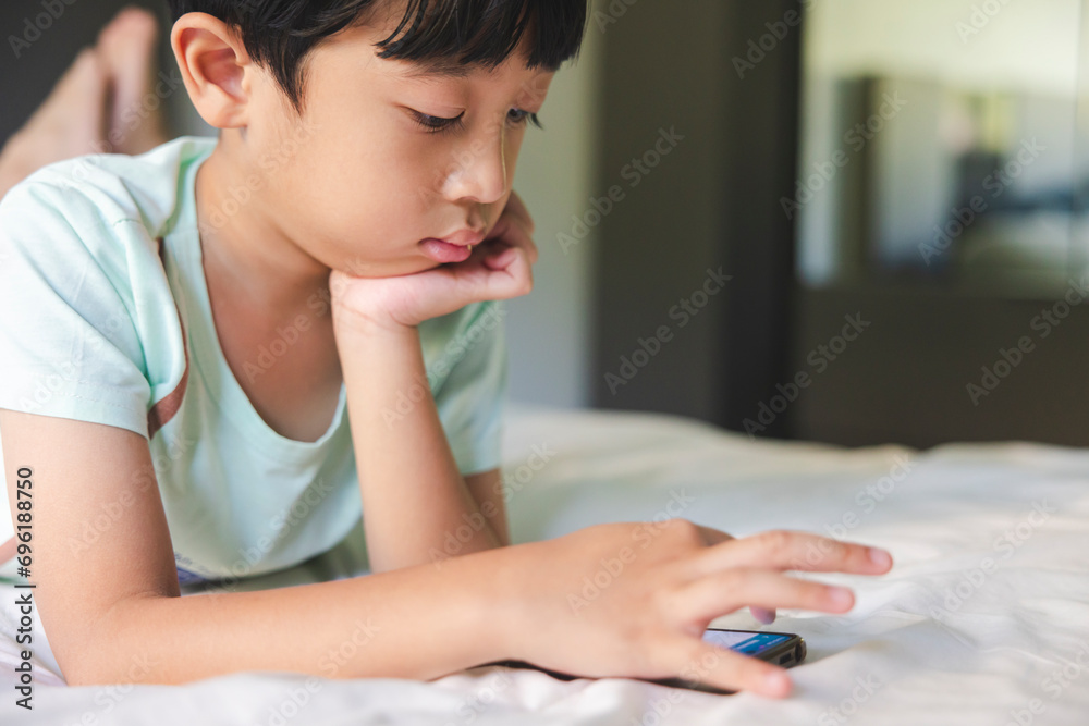 Close up portrait of Asian boy with black bangs, black eyes with a smiling face wearing a light green shirt lying on the bed in his house playing with his smart phone. Education concept. Close up portrait of Asian boy with black bangs, black eyes with a smiling face wearing a light green shirt lying on the bed in his house playing with his smart phone. Education concept.