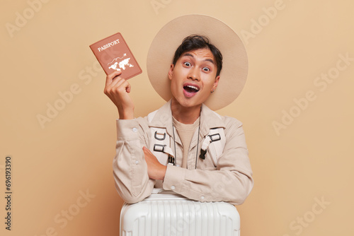 Young man dressed in beige shirt and broadbrim hat shows his passport with wide smile, journey time concept, copy space photo