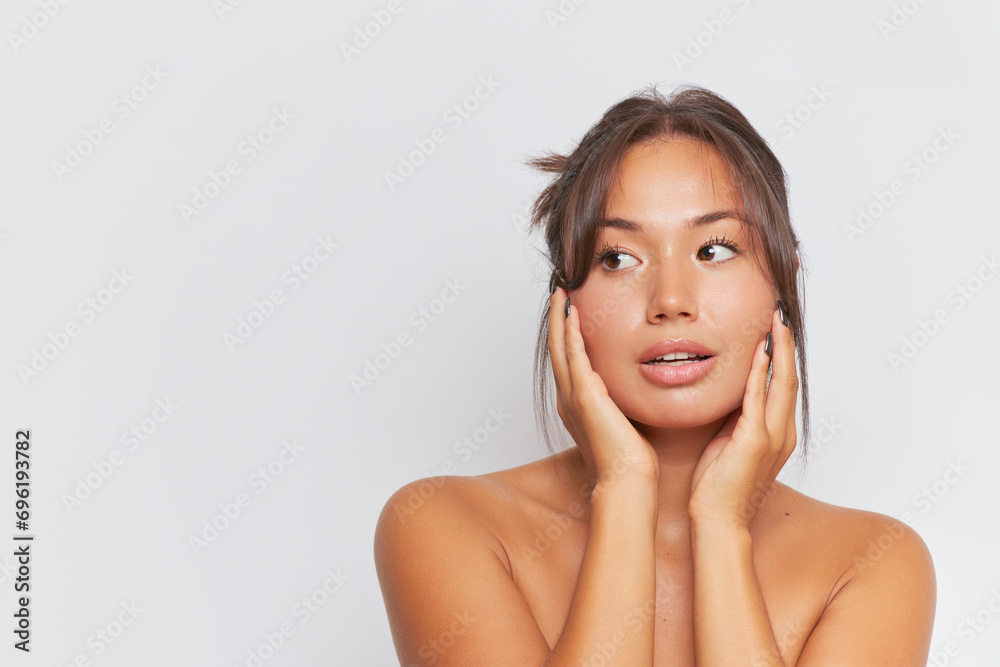 Beautiful woman with naked shoulders pressing hands to her face looking aside, skincare product concept, copy space