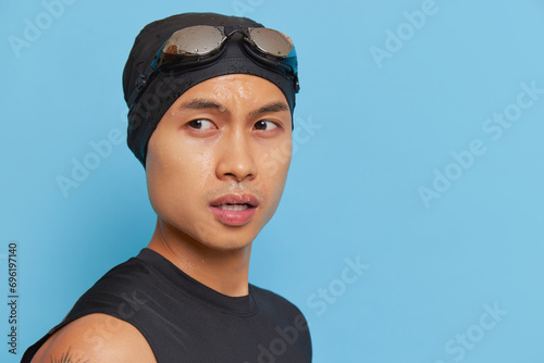 Portrait of sportive young man in swimming cap with goggles standing on blue background looking aside, sport life concept, copy space © South House Studio