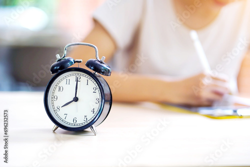 Selective focus of alarm clock on table near young female student in casual cloth doing homework at her school. concept of time management. photo
