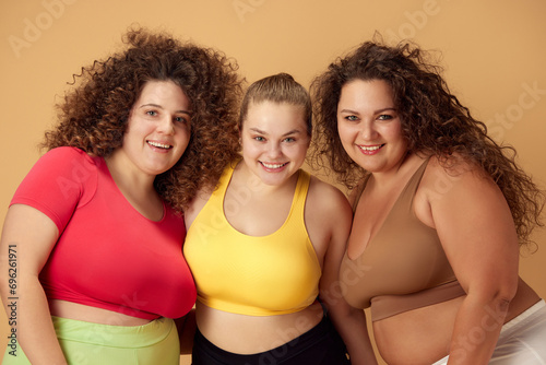 Portrait of three beautiful, smiling, overweigh women standing in sportswear over beige studio background. Concept of sport, body-positivity, weight loss, body and health care