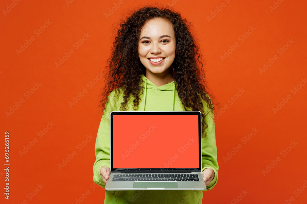 Naklejka premium Smiling fun young IT woman of African American ethnicity wear green hoody casual clothes hold use work on blank screen laptop pc computer isolated on plain red orange background. Lifestyle concept.