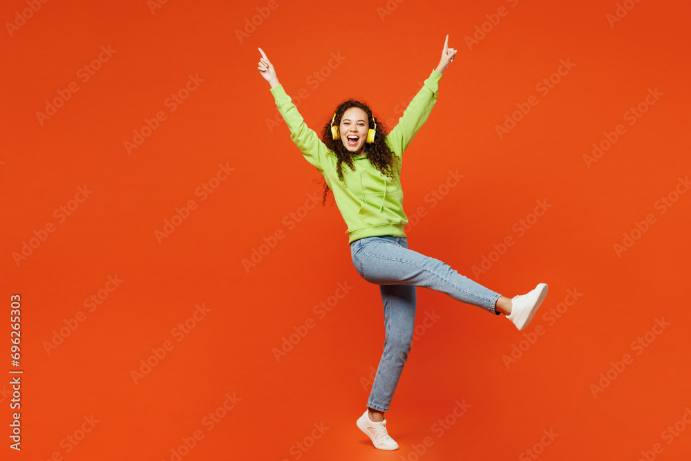 Full body happy young woman of African American ethnicity she wear green hoody casual clothes listen to music in headphones raise up hands isolated on plain red orange background. Lifestyle concept.