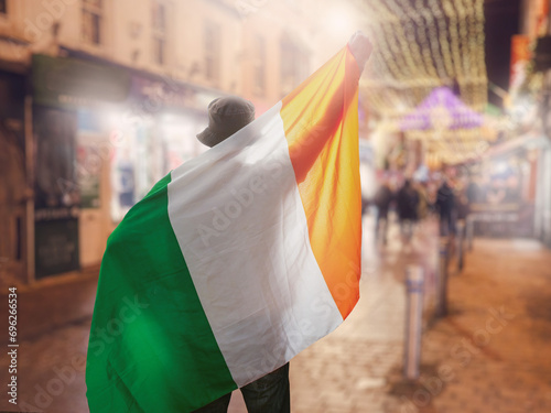 Man holding National flag of Ireland high and on his back, illuminated city street and traffic out of focus in the background. Sport fan in town celebrating concept.