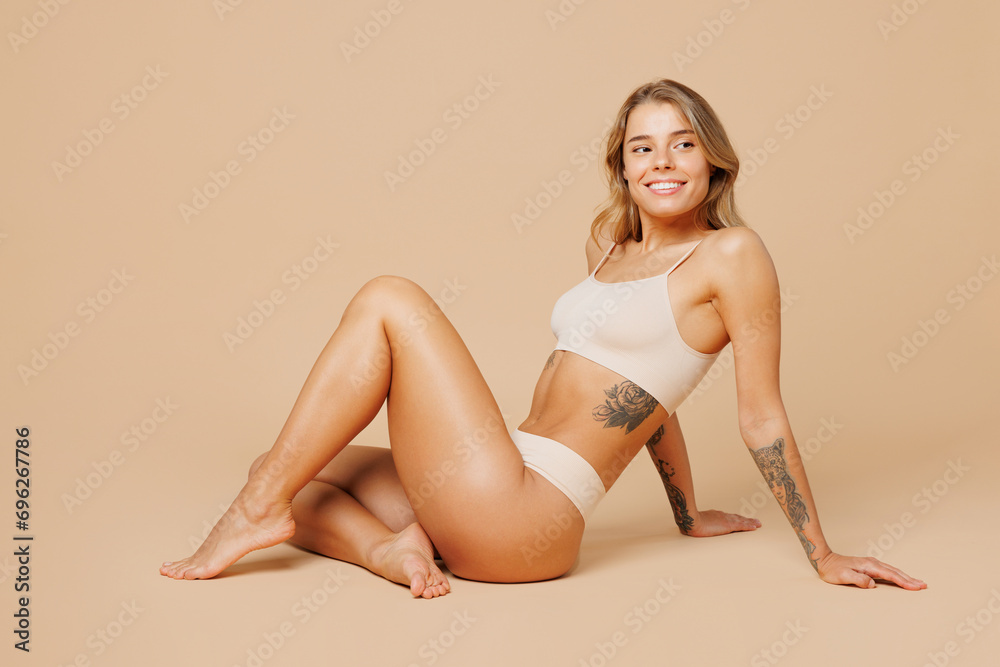 Full body side view happy fun young nice lady woman with slim body perfect skin wear nude top bra lingerie sitting looking aside isolated on plain pastel beige background. Lifestyle diet fit concept. - obrazy, fototapety, plakaty 