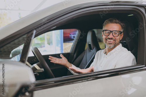 Close up smiling fun adult man customer male buyer client wear shirt sit in car spread hands choose auto want to buy new automobile in showroom vehicle salon dealership store motor show Sales concept