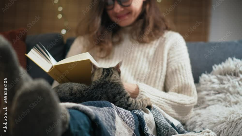 A young Caucasian woman reading a book sitting on the couch with her cute grey cat at home on a winter evening