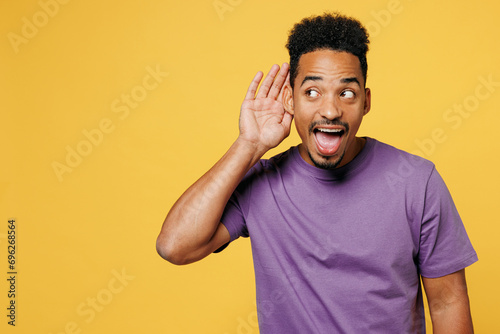 Young nosy man of African American ethnicity he wears purple t-shirt casual clothes try to hear you overhear listening intently isolated on plain yellow background studio portrait. Lifestyle concept. © ViDi Studio