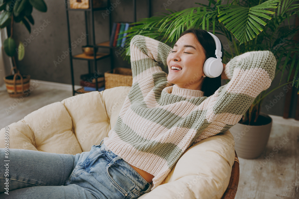 Obraz premium Side view young fun woman she wear casual clothes listen music in headphones sits on armchair stay at home hotel flat rest relax spend free spare time in grey living room indoor People lounge concept
