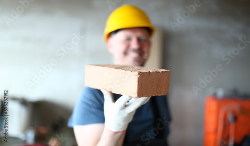 Focus on serious brick in male hand. Worker in hardhat presenting red block to camera with big smile. Joyful constructor in yellow helmet looking with happiness on face. Building concept photo