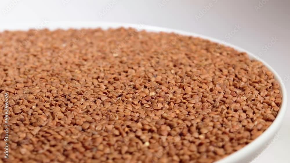 Dry Uncooked Brown Buckwheat Groats on White Plate Rotating against White Background. Raw Large Buckwheat Grains. Russian, Ukrainian and Belarusian Culture Dry Uncooked Brown Buckwheat Groats on White Plate Rotating against White Background. Raw Large Buckwheat Grains. Russian, Ukrainian and Belarusian Culture