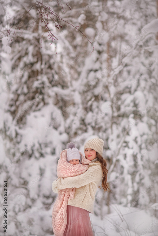 Fototapeta premium Winter. Snow. Mom and baby in the winter forest. Portrait. Mom gently holds her daughter in her arms. The baby is wrapped in a warm blanket. Trees in the snow. Family. Mom and daughter. Cold. Snowy