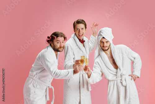 Happy New Year. Positive young men, friends in bathrobes after shower, clinking champagne glasses against pink background. Concept of leisure activity, fun, bachelor party, friendship, spa