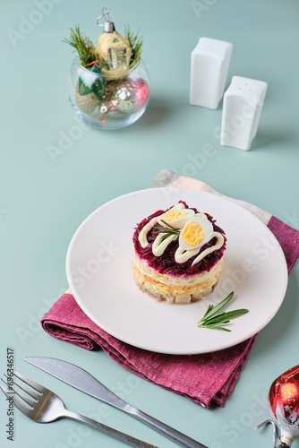 Portioned herring salad under a fur coat, herring under layers of boiled vegetables, eggs, smeared with mayonnaise, on a white plate on a green plain background.