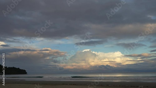 Early morning on a tropical beach. The clouds in the blue sky are highlighted in gold and pink. The waves of the turquoise ocean roll towards the shore, foaming. Glare on the water. Malaysia. Borneo. 