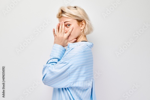 Tableau sur toile Portrait of young blonde woman covering eyes with hand, peeking through fingers isolated on white studio background