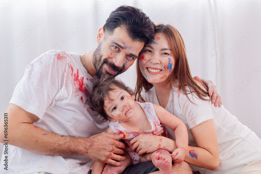 Naughty adorable family with little toddler having fun laughing playing together at home, Hispanic father and Asian mother spend great time with daughter painting faces in relaxation holiday