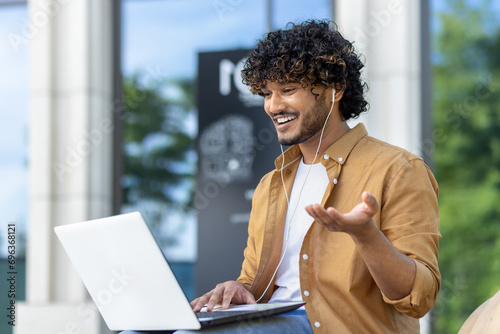 Muslim young man sitting on a bench on a city street wearing headphones and smilingly talking on a video call on a laptop while gesturing with his hands photo