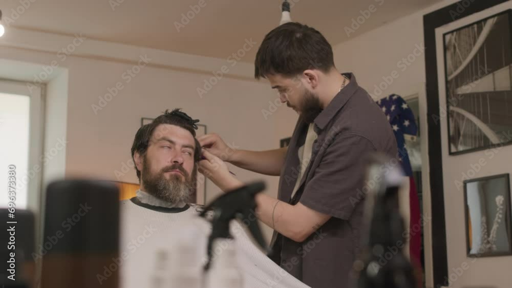 Bearded man in hairdressing cape sitting in barbershop while young barber cutting his hair, USA flag and black-and-white photos on wall in background Bearded man in hairdressing cape sitting in barbershop while young barber cutting his hair, USA flag and black-and-white photos on wall in background
