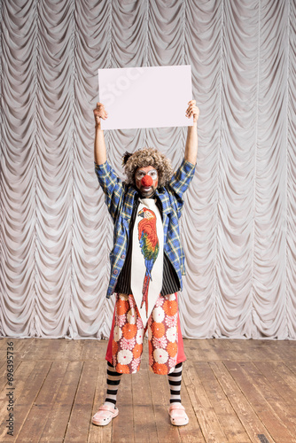 A funny clown holds an empty cardboard box on which you can paste text. A clown on the background of beige stage scenes