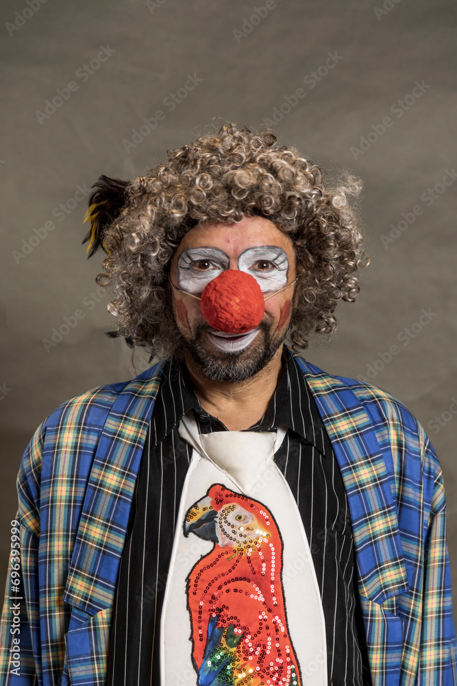 A cheerful curly-haired clown grimaces. A close-up portrait against a background of beige wings. The clown in the parrot tie A cheerful curly-haired clown grimaces. A close-up portrait against a background of beige wings. The clown in the parrot tie