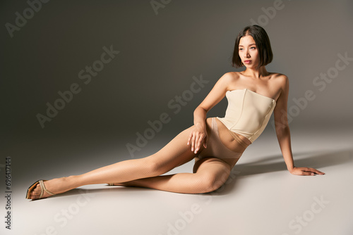beautiful asian model in corset and beige fishnet tights sitting while posing on grey background © LIGHTFIELD STUDIOS