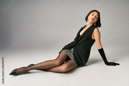 chic and beautiful young asian woman in elegant outfit with gloves sitting on grey background